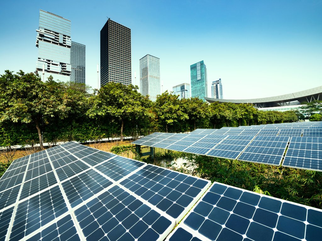 Fotografía de un edificio parte de la construcción sustentable, con paneles solares instalados en el techo. Los paneles solares son de color azul oscuro y están dispuestos en filas ordenadas. El edificio es de color blanco y tiene varios pisos. Al fondo se observa un cielo azul claro con algunas nubes blancas